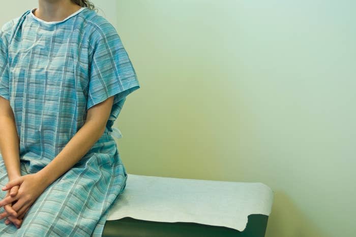 A person sitting on a medical examination table, dressed in a hospital gown, with hands folded on their lap