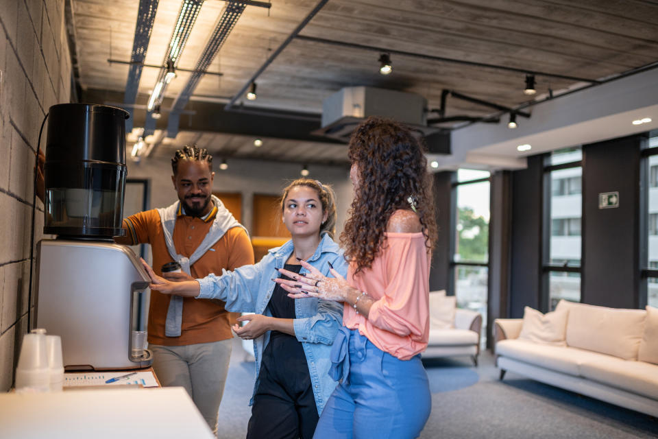 Three colleagues, a man and two women, are discussing at a coffee machine in an office lounge. A woman gestures with her hands