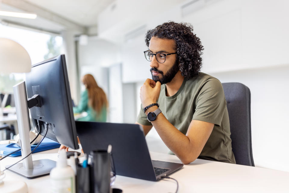 A person with glasses and curly hair sits at a desk and looks intently at a computer screen in a modern office environment, with another person blurred in the background