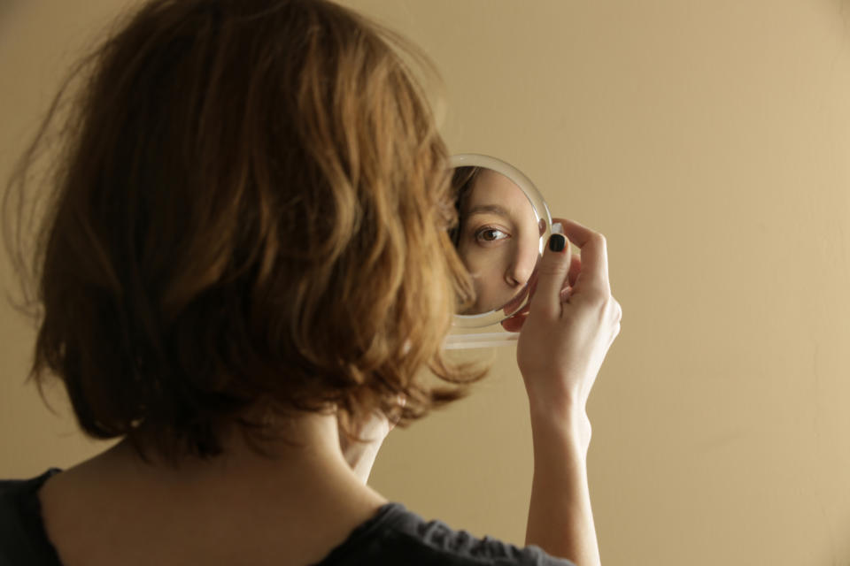 A person with shoulder-length hair looks at his reflection in a hand mirror, with only one eye visible