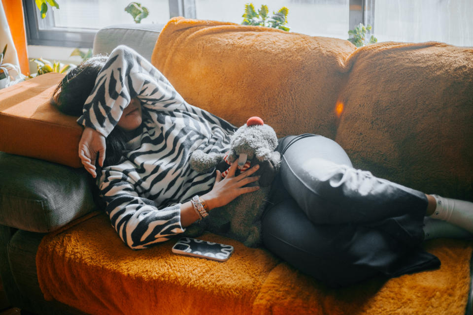 Person lying on a couch wearing a zebra print sweater, holding a stuffed animal and covering his face with one arm