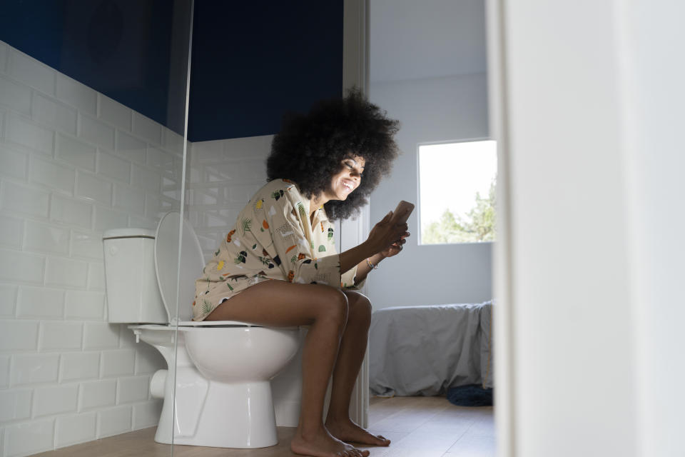 Person sitting on a toilet, smiling while looking at a phone. They are wearing patterned loungewear. The bathroom has a window that lets in natural light