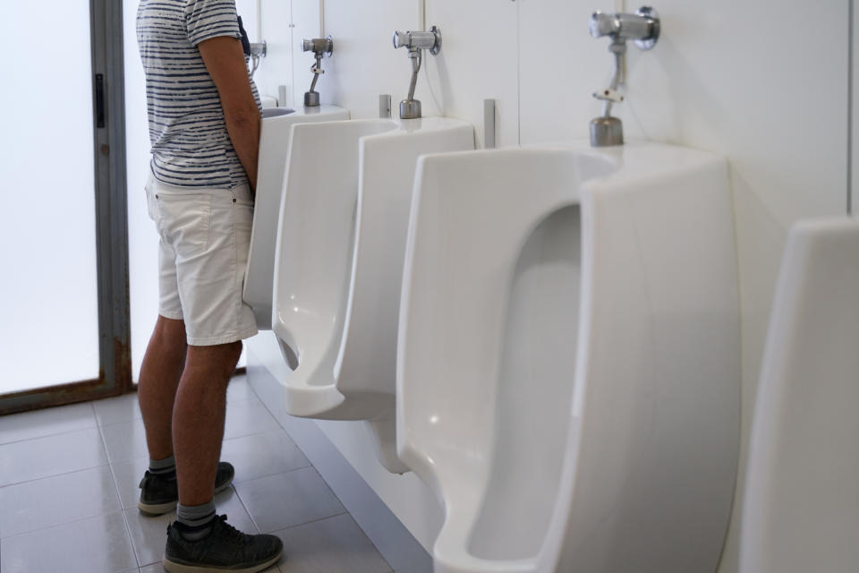 A person in a striped shirt and shorts stands in front of a row of urinals in a bathroom