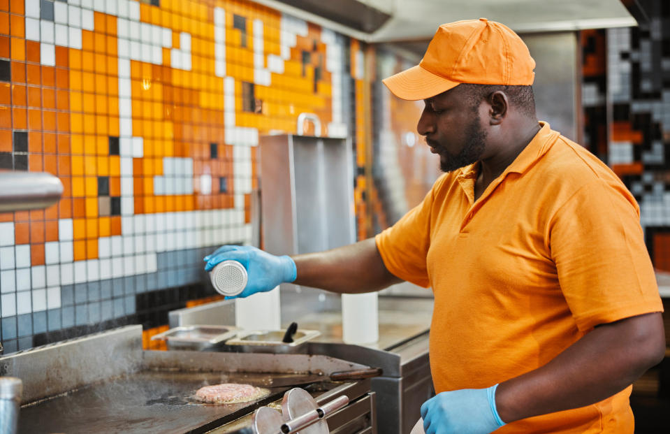 A man in an orange polo shirt and cap grills a burger and spices it in a professional kitchen. He wears blue gloves for hygiene