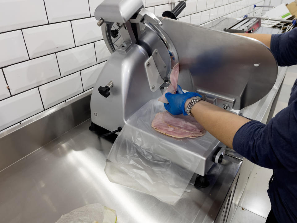 Person in long sleeve shirt and gloves cuts meats on meat machine in commercial kitchen