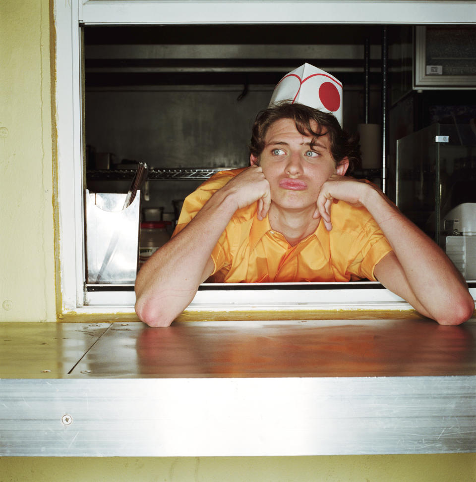 A person in a fast food worker uniform and hat leans on a counter, looking bored. They are standing in a service window, their chin resting on their hands