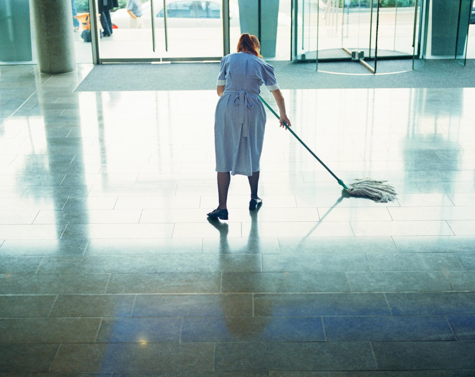 A uniformed janitor mops a large tiled floor in the lobby of an office building, with sliding doors and windows in the background