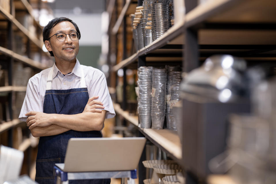 A man wearing glasses and an apron stands with folded arms in a warehouse aisle, smiling and looking to the side, with metal goods and a laptop in the foreground