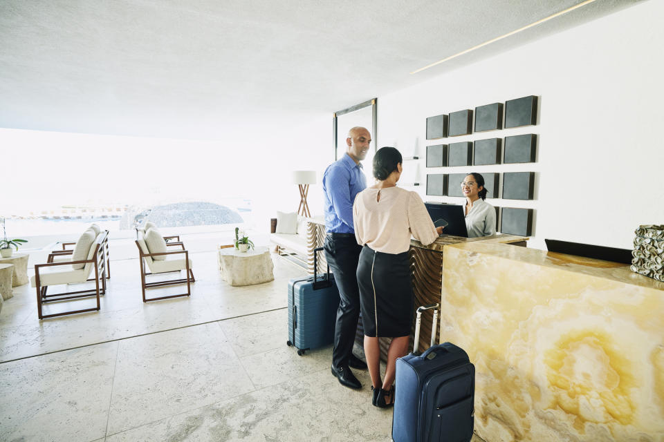 Two people with luggage check in at a hotel reception, assisted by a receptionist