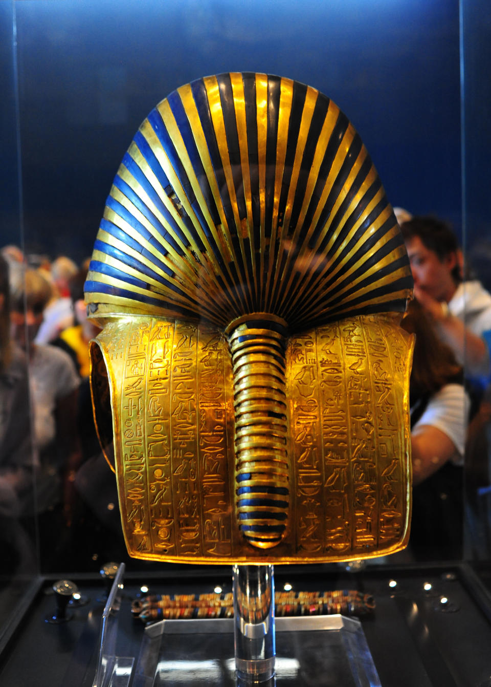 Golden ancient Egyptian pharaoh mask with intricate hieroglyphs on display, viewed from behind with people watching in the background