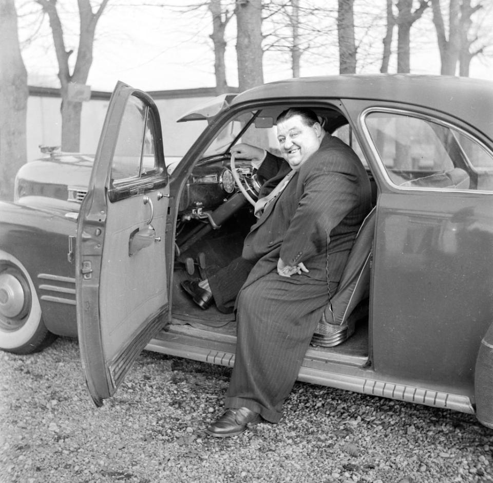 A man in a suit sits in the driver's seat of an open car door and smiles at the camera