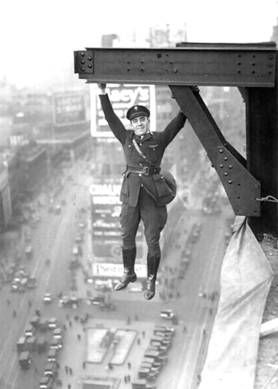 A man in uniform hangs from a steel beam high above a busy city street in an old photo