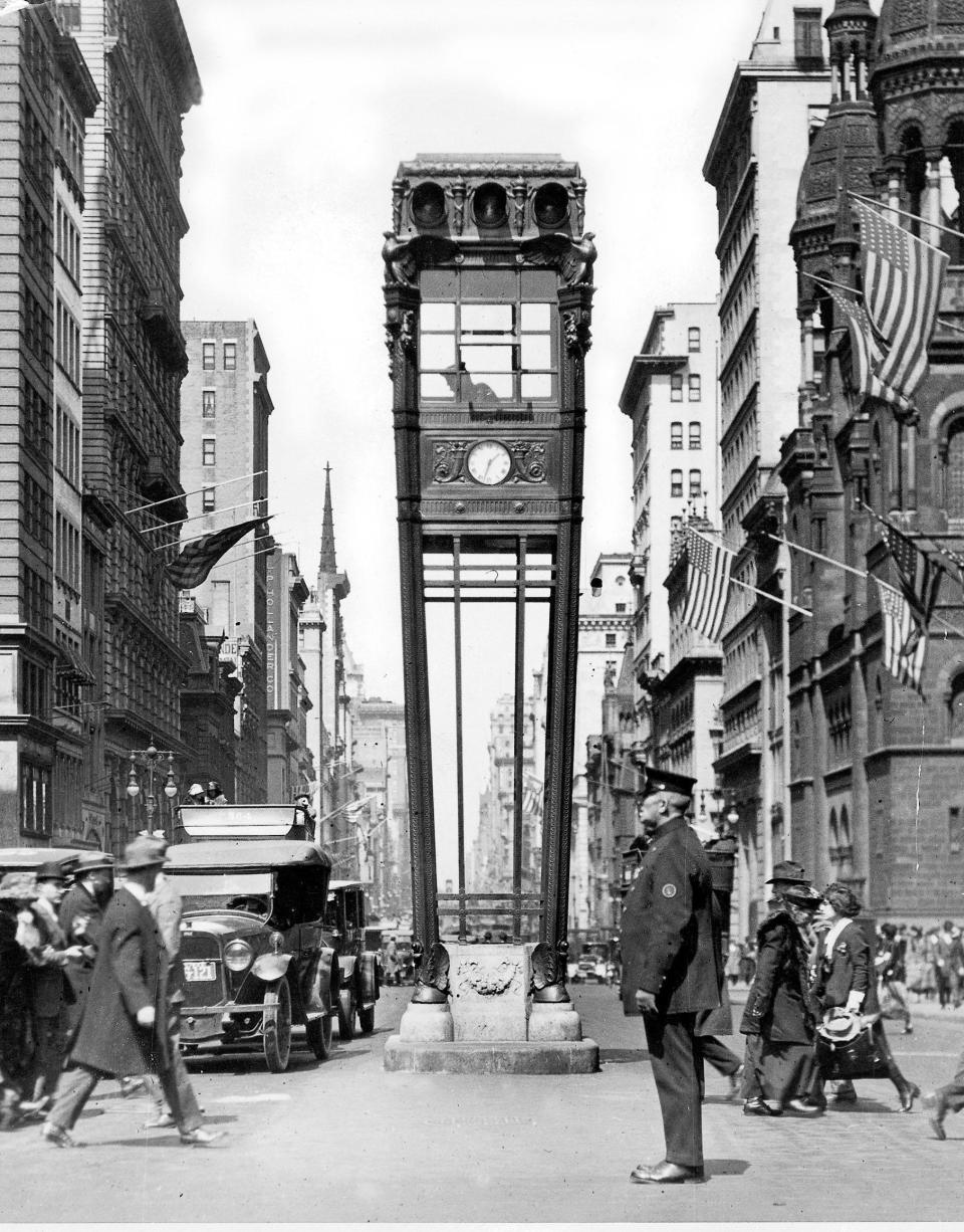 Vintage photo of New York City's 5th Avenue with a tall clock structure in the middle of the street. The street is full of people and old cars