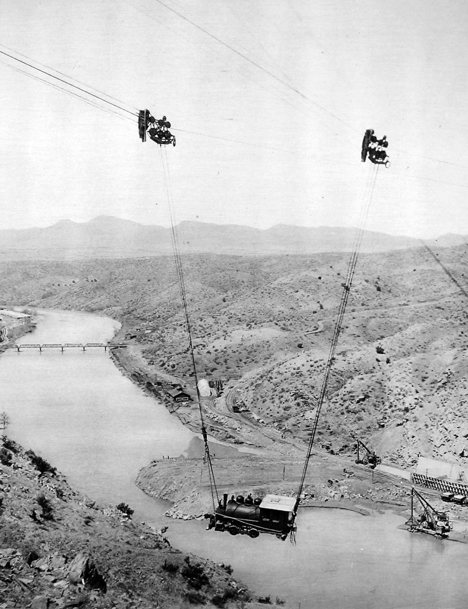 Historic photo of construction workers hanging from cables over a river gorge during the construction of the Roosevelt Dam in Arizona, early 20th century