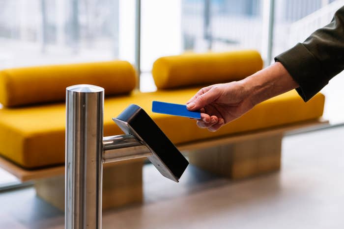 A person uses a blue key card to access a modern revolving gate in an office lobby with yellow seating in the background