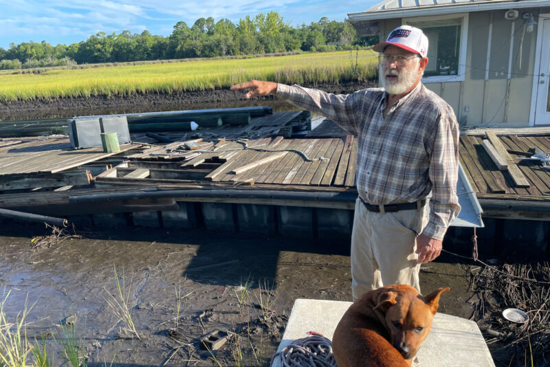 An elderly man points to the rising tide while standing on a jetty.