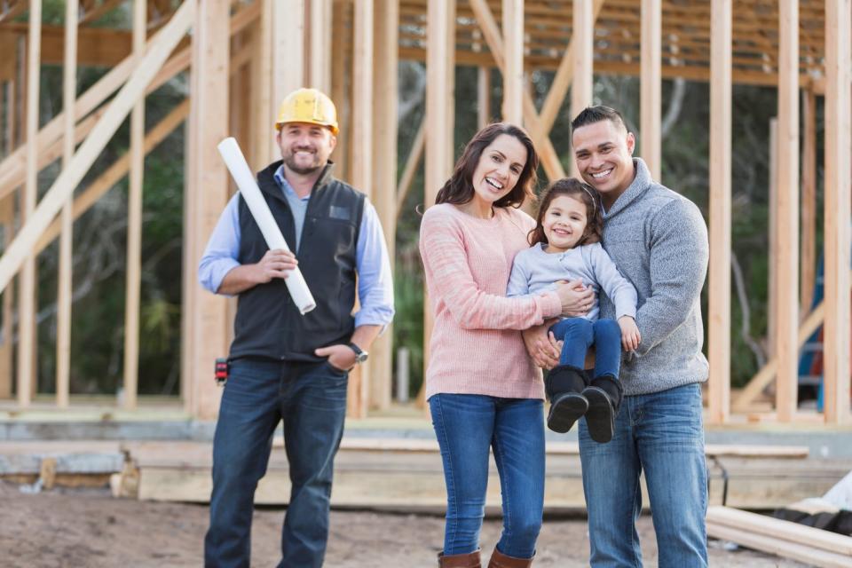 A family and a construction worker laugh as they stand in front of their future home, which is still in the early stages of construction.