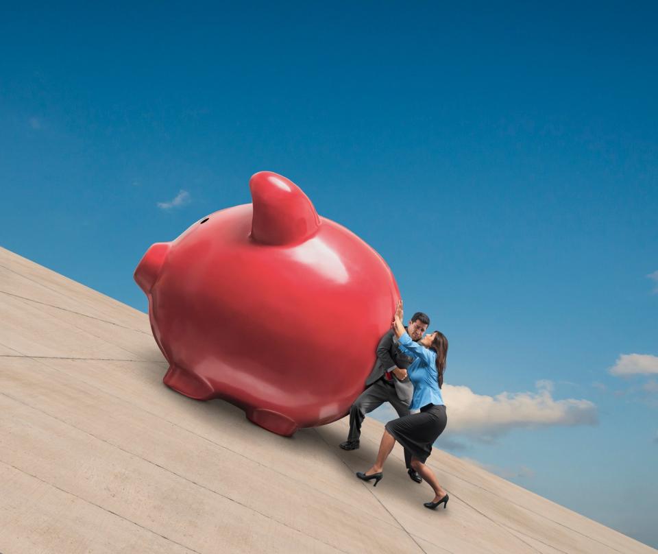 Two people push a giant piggy bank up a slope.