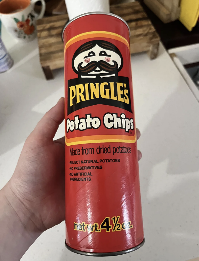 A hand holds a can of Pringle's Potato Chips. The label reads: "Made from dried potatoes. Select natural potatoes. No preservatives. No artificial ingredients."