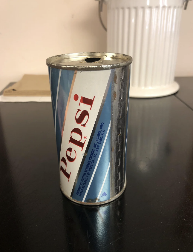 A vintage Pepsi can with a pop-top opening sits on a dark table. A white, ribbed container is faintly visible in the background