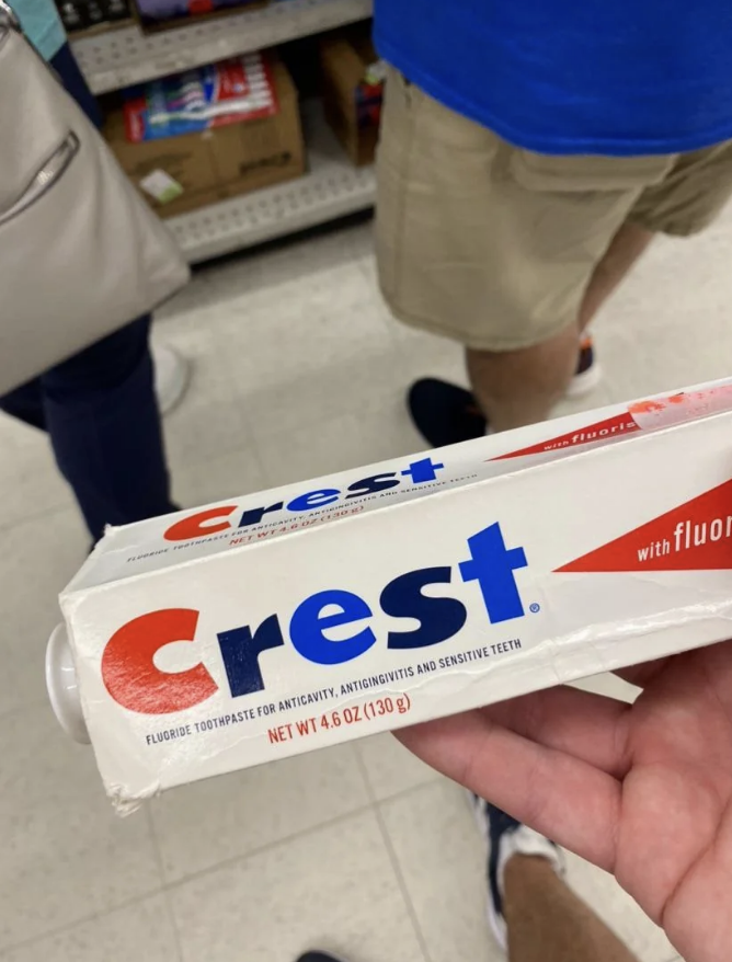 A person holds a 4.6 oz tube of Crest toothpaste in a store. The text on the toothpaste reads: "Fluoride toothpaste against cavities, anti-gingivitis and sensitive teeth."