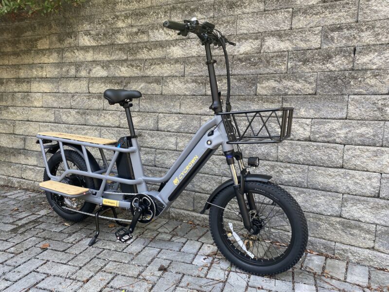 Image of a gray cargo bike with a long tail, high handlebars and a basket at the front, and a wooden deck and footrests at the loading area.