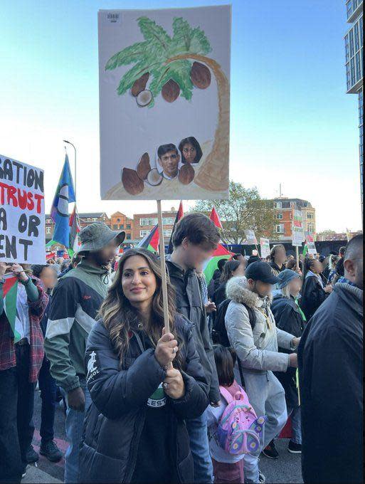 Marieha Hussain pictured at the march holding a sign with a drawing of a palm tree with coconuts falling from it. Photos of Rishi Sunak and Suella Braverman are pasted onto two of the coconuts. A crowd of protesters can be seen in the background of the image. 