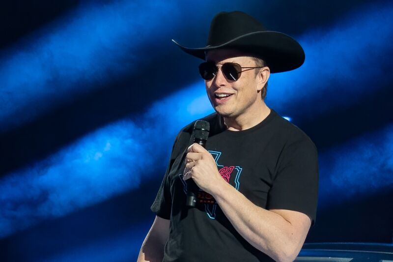 Elon Musk speaks at an event wearing a cowboy hat, sunglasses and a T-shirt.