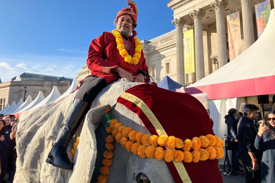 A man with a red turban and yellow garland wearing a comical costume of an elephant, making it look like he is sitting on it