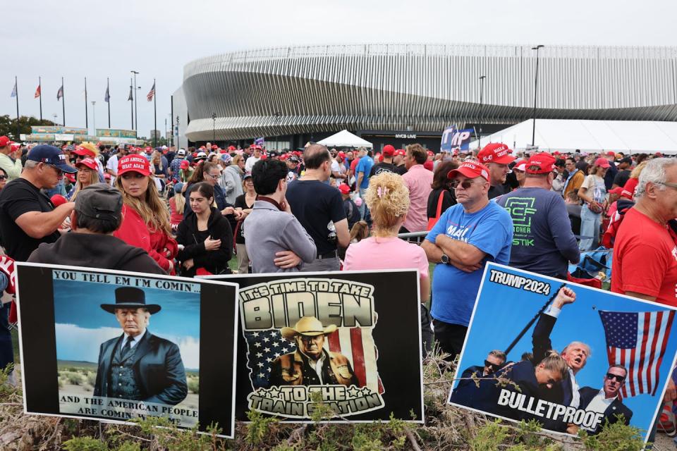 On a gray day, a crowd of people stand outside, in front of three large signs with a man dressed in a Wild West costume, with the words 'Just tell me I'm coming', and a poster of a man surrounded by men in suits while raising his fist