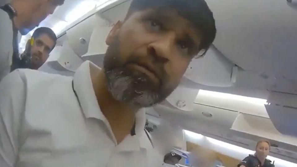 A man with dark hair and a dark and gray beard looking just above the camera. He is wearing a white polo shirt and sitting on a plane