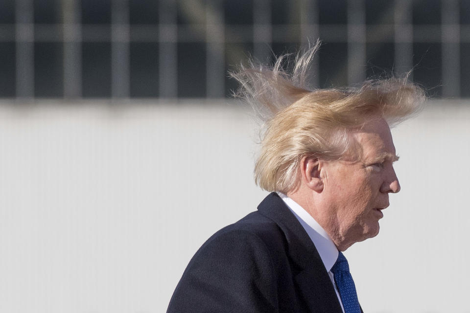 A person with wind-swept hair wearing a dark suit and tie walks outside in front of a building with reflective windows