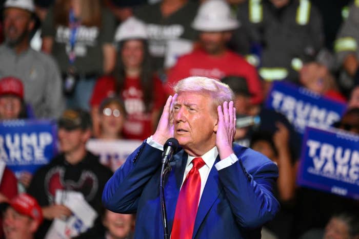 Person in suit and red tie speaks into a microphone at a busy event, with people holding signs in the background