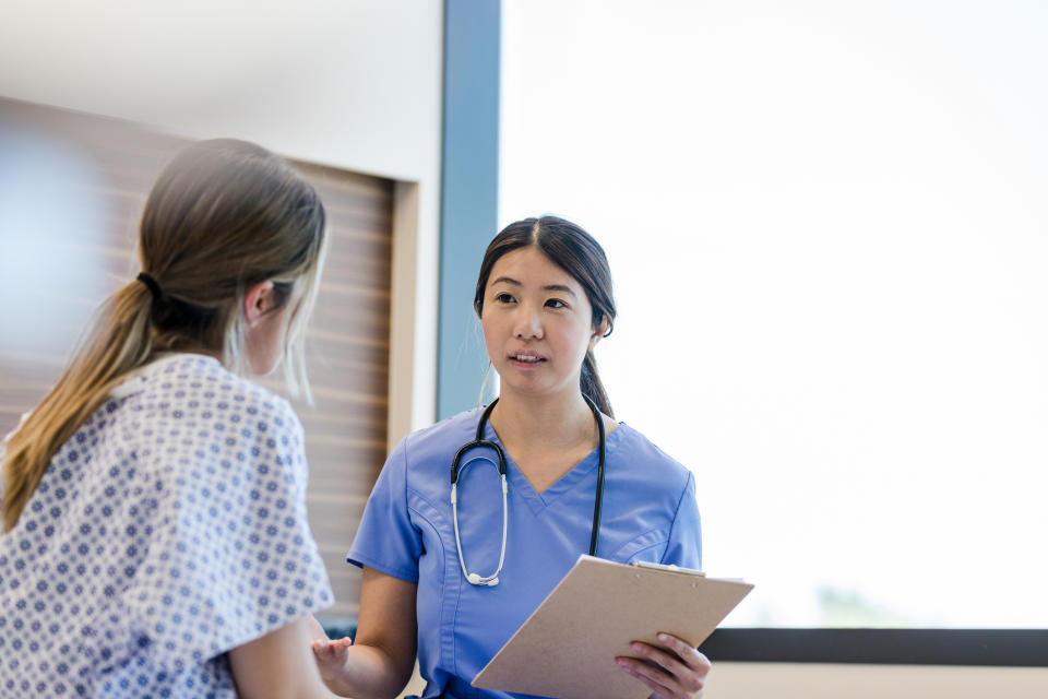 A nurse in scrubs holds a clipboard and talks to a patient wearing a hospital gown in a medical setting