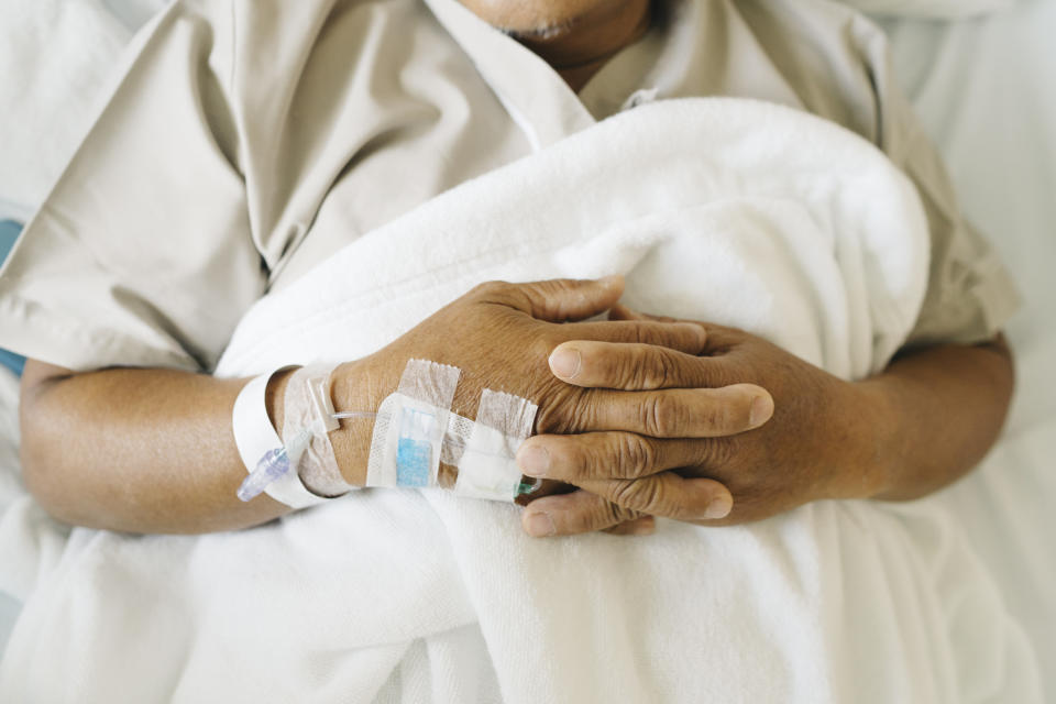 Person in hospital bed with an IV in hand, both hands resting on a blanket