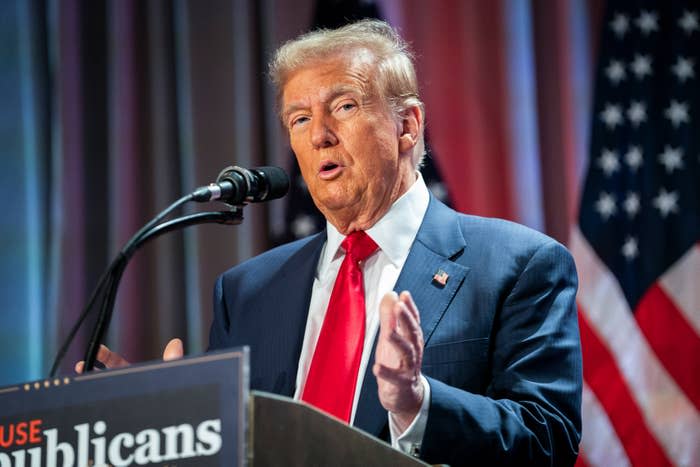 A person in a suit and tie speaks into a microphone and gestures with his hands at an event. American flags are visible in the background