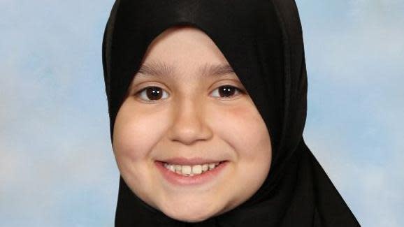 A photo of Sara Sharif smiling and wearing a black headscarf. She is looking at the camera and in front of a blue background