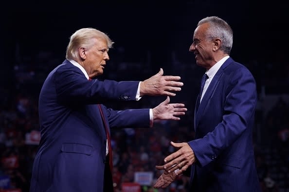 Two men in suits greet each other warmly at a podium and extend their arms as they walk in for a handshake or hug