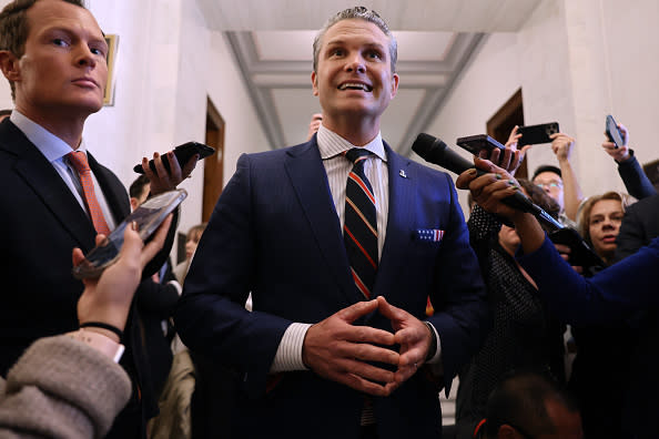 A man in a suit and striped tie is surrounded by journalists holding microphones and phones in a hallway