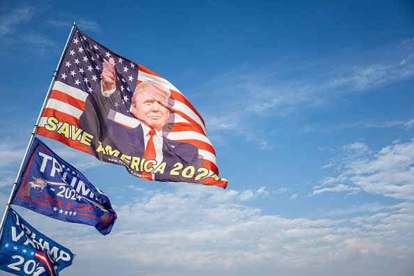 Flags with a political figure and "Save America 2024" text wave against a blue sky