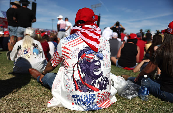 Person sitting on the grass during an outdoor event, wearing a shirt with an image of a man in a suit, patriotic accessories and the word "TRUMP."