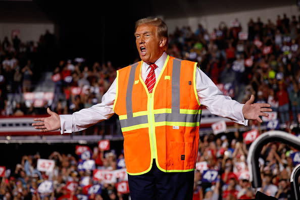 Person on stage, wearing a reflective safety vest, with arms outstretched, speaking to a cheering crowd at an event