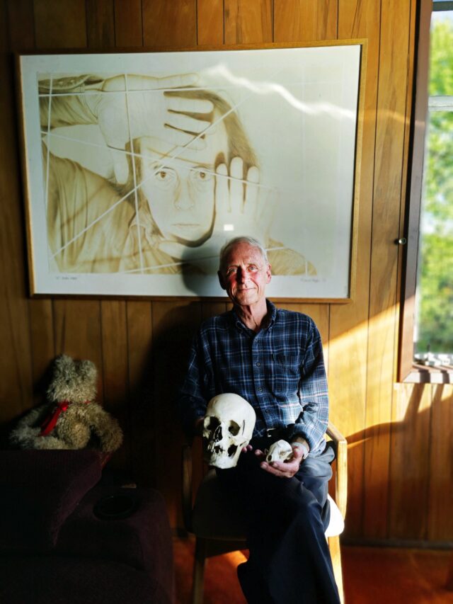 older white man in plaid shirt sitting under artwork with a skull on his lap