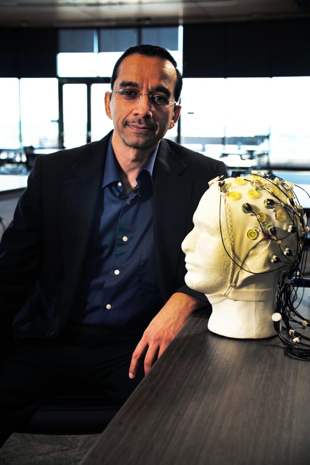 Rajesh Rao sits next to a plaster head and models a brain-computer interface device with a skull cap