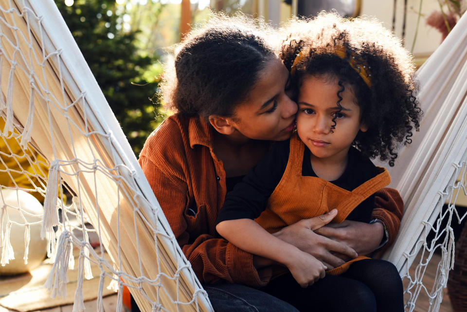 A woman lovingly kisses a young child on the cheek as she sits outdoors in a hammock. Both wear cozy, casual outfits, which suggest a relaxing moment