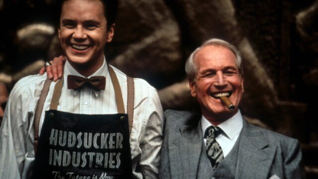 young man in mailroom apron beams and stands next to an older man in a suit with a cigar clenched between his teeth