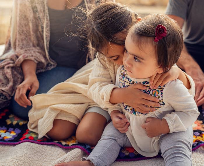 A young girl lovingly embraces a toddler on a blanket. They are both dressed casually