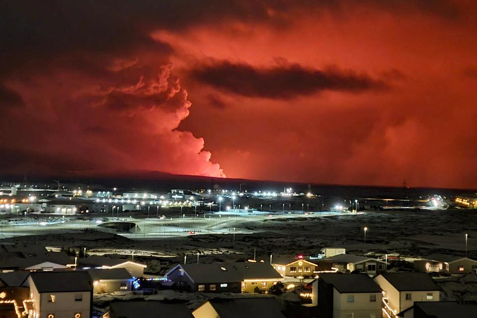 Houses in the village of Hafnarfjordur are seen as smoke billows in the distance as lava turns the night sky orange after a volcanic eruption on the Reykjanes Peninsula, western Iceland on December 18, 2023. A volcanic eruption began Monday evening in Iceland, south of the capital Reykjavik, after an earthquake swarm, the Icelandic Meteorological Office reported. (Photo by Oskar Grimur Kristjansson / AFP) (Photo by OSKAR GRIMUR KRISTJANSSON/AFP via Getty Images)