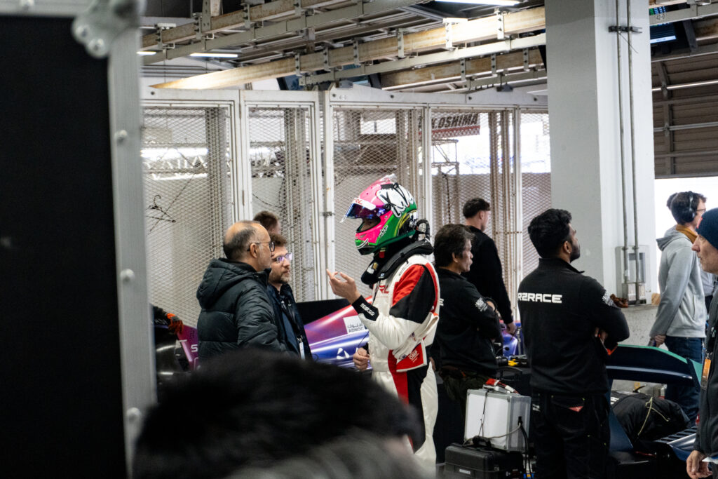 A driver and some engineers stand in a garage on a race track