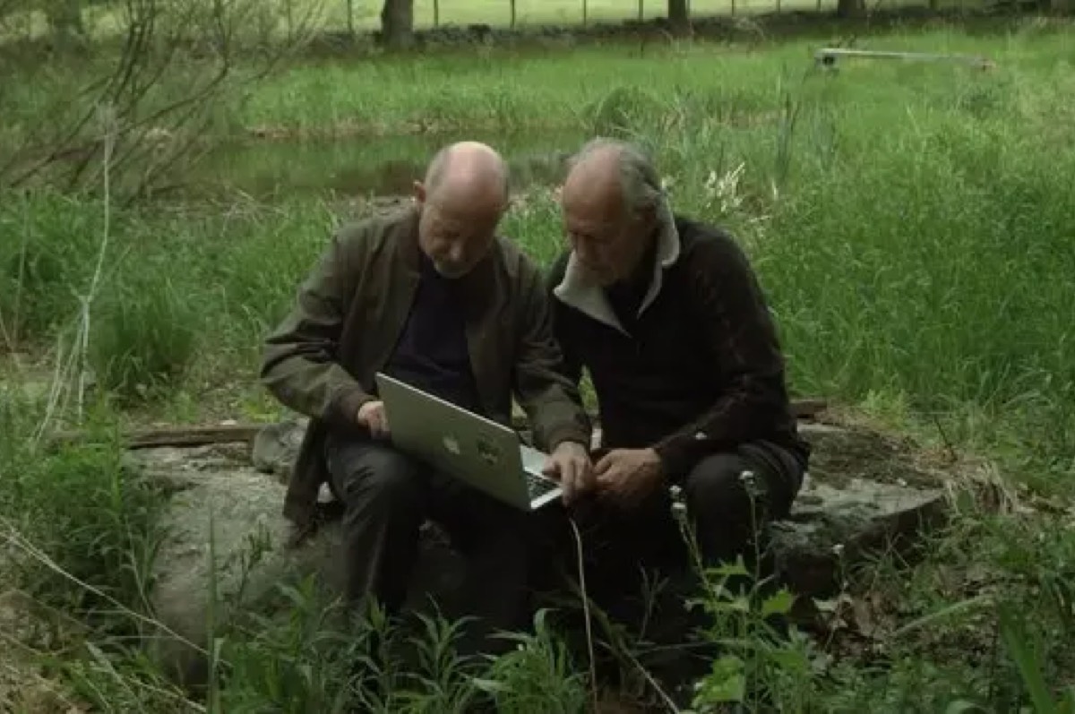 Werner Herzog and Rafael Yuste in a field, sitting on a tree trunk while looking at a laptop screen